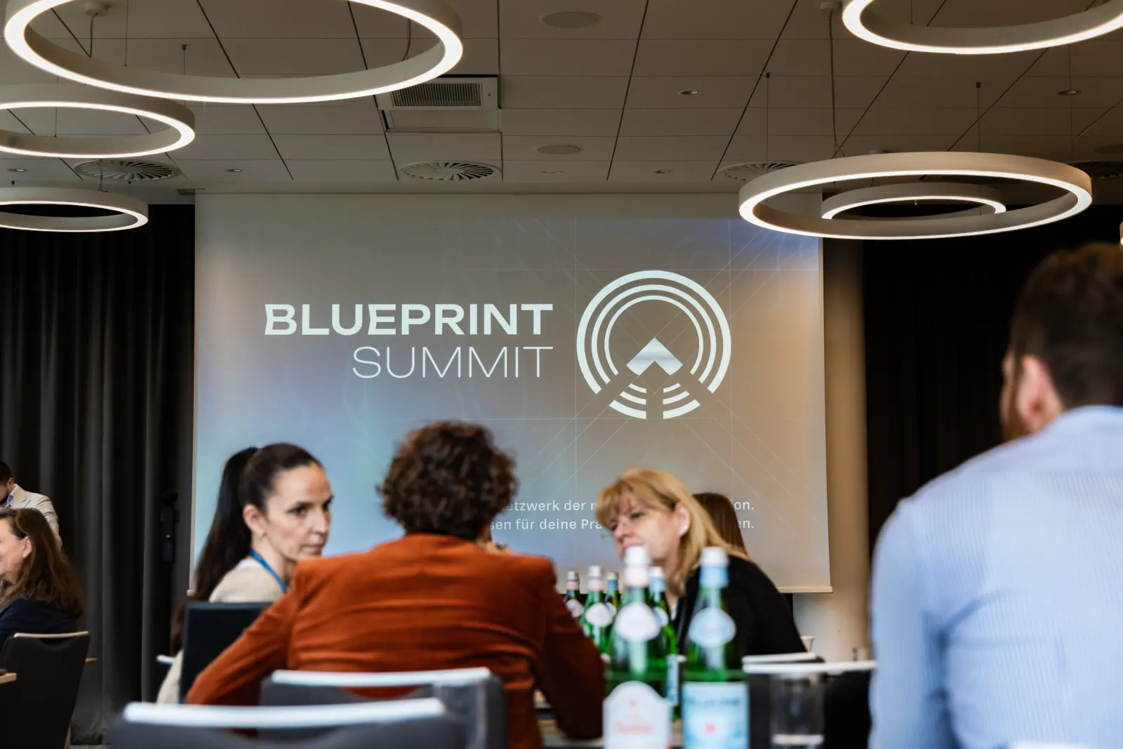 Das Logo des Blueprint Summit auf der Leinwand, davor Teilnehmende im Gespräch